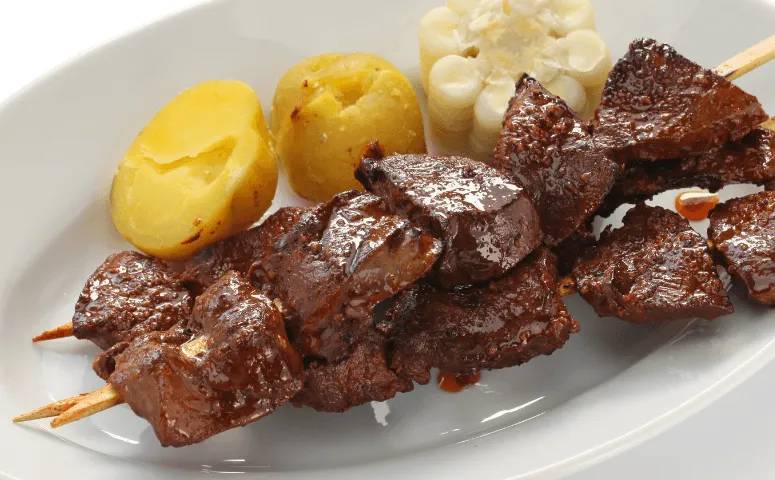 Brochetas de anticuchos de corazon de res a la parrilla con papas en un plato blanco. 